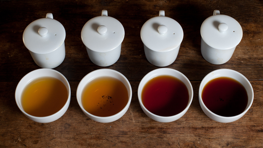 Four cups of brewed tea in different colours beside matching white teapots on a wooden table