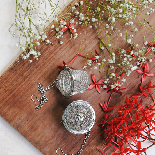 Silver mesh tea infusers on a wooden board with red and white flowers