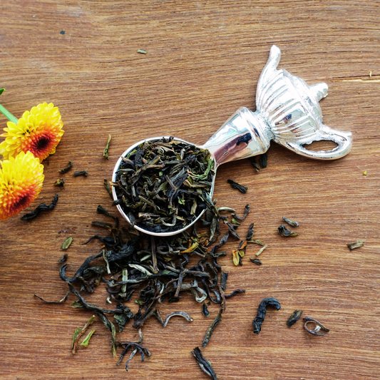 Silver tea measuring spoon with loose leaf tea and yellow flowers on wooden surface